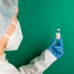 A healthcare worker wearing gloves and a mask holds a COVID-19 vaccine vial against a green background.